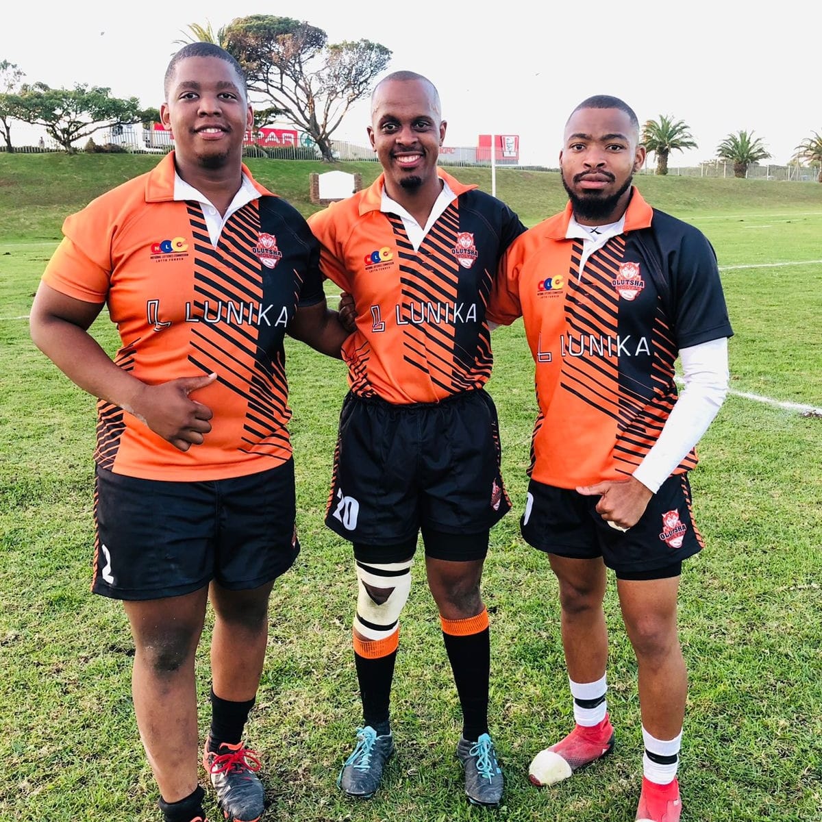 Three Olutsha players in orange and black club kit standing together on the field